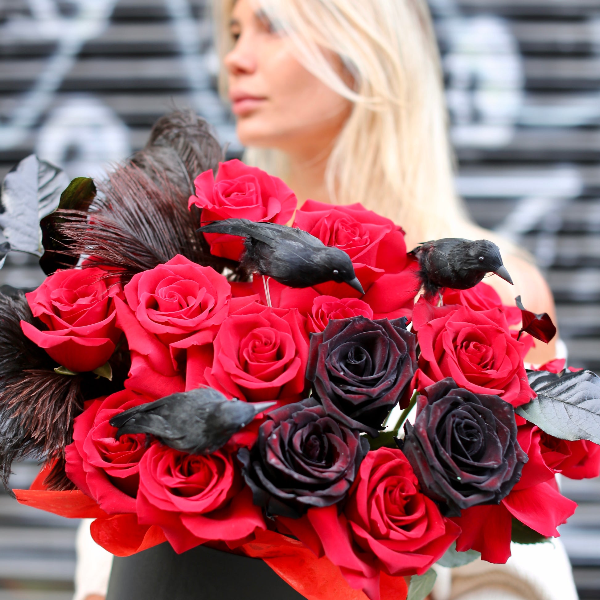 Elegant red and black rose arrangement styled with feathers and seasonal accents, ideal for Halloween gifting